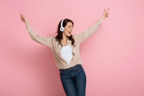 Cheerful woman pointing with fingers while listening music in headphones on pink background — Stock Photo