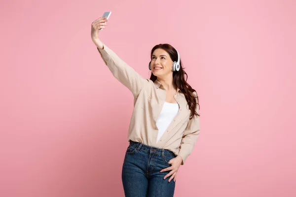 Smiling woman in headphones taking selfie with smartphone on pink background, concept of body positive — Stock Photo