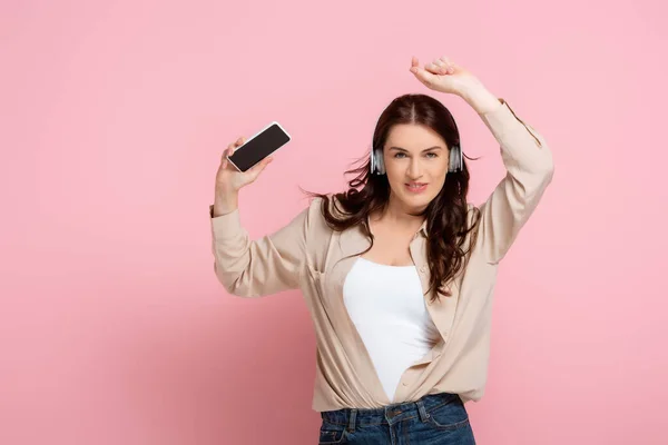 Femme positive dans les écouteurs tenant smartphone et regardant la caméra sur fond rose — Stock Photo