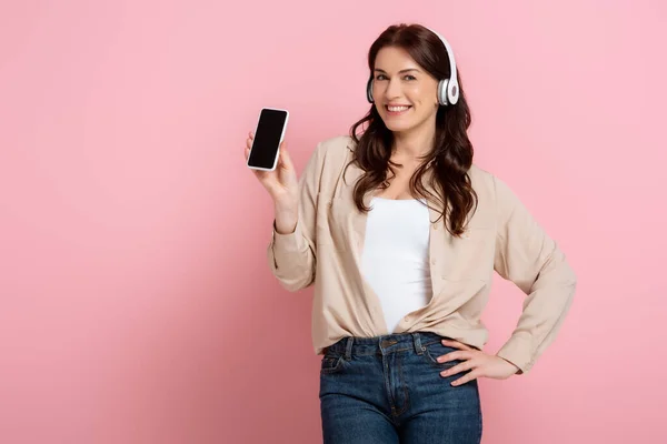 Hermosa mujer con auriculares sosteniendo el teléfono inteligente y sonriendo a la cámara sobre fondo rosa - foto de stock