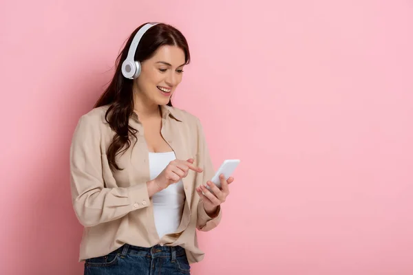 Mujer positiva en auriculares apuntando con el dedo al teléfono inteligente sobre fondo rosa — Stock Photo
