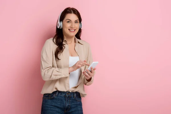 Mujer atractiva sonriendo a la cámara mientras escucha música en los auriculares y utilizando el teléfono inteligente sobre fondo rosa — Stock Photo