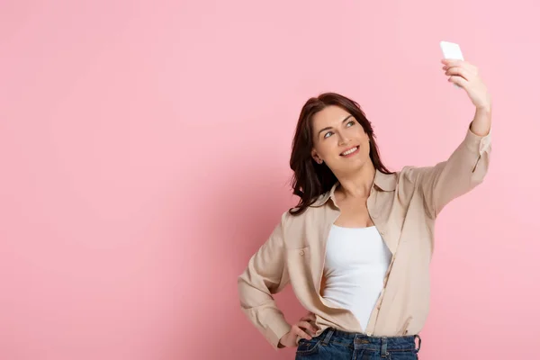 Mulher morena sorridente posando enquanto toma selfie no smartphone no fundo rosa — Fotografia de Stock