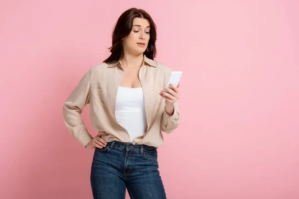 Skeptical woman with hand on hip using smartphone on pink background, concept of body positive — Stock Photo