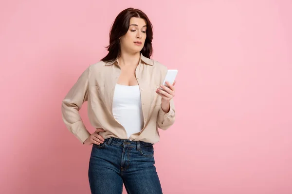 Mulher cética com a mão no quadril usando smartphone no fundo rosa, conceito de corpo positivo — Fotografia de Stock