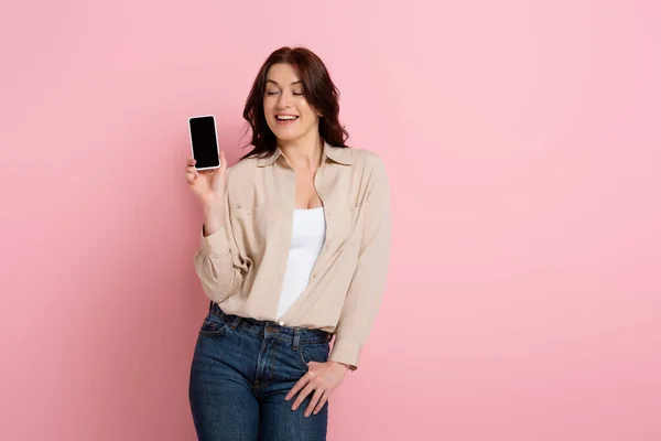 Hermosa mujer sonriente mostrando teléfono inteligente con pantalla en blanco sobre fondo rosa, concepto de cuerpo positivo - foto de stock