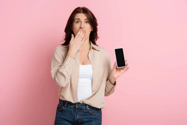 Shocked woman covering mouth with hand and showing smartphone on pink background, concept of body positive — Stock Photo