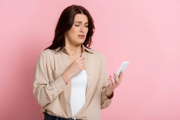 Donna preoccupata che tiene smartphone su sfondo rosa, concetto di corpo positivo — Foto stock