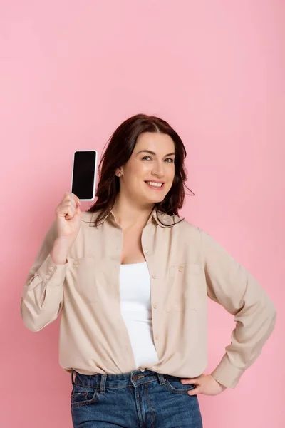 Bella donna sorridente con mano sul fianco mostrando smartphone con schermo bianco isolato su rosa — Foto stock