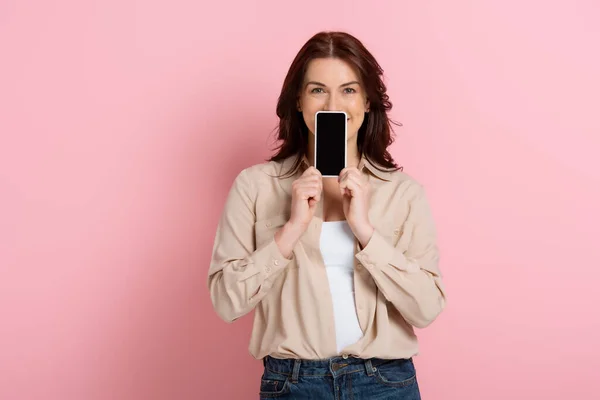 Brunette woman showing smartphone with blank screen near face on pink background — Stock Photo