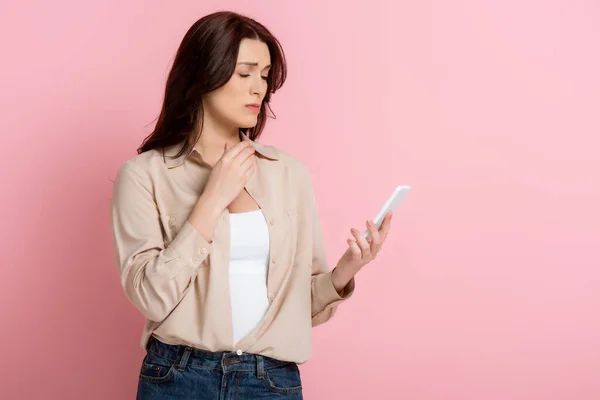 Donna bruna triste utilizzando smartphone su sfondo rosa, concetto di corpo positivo — Foto stock