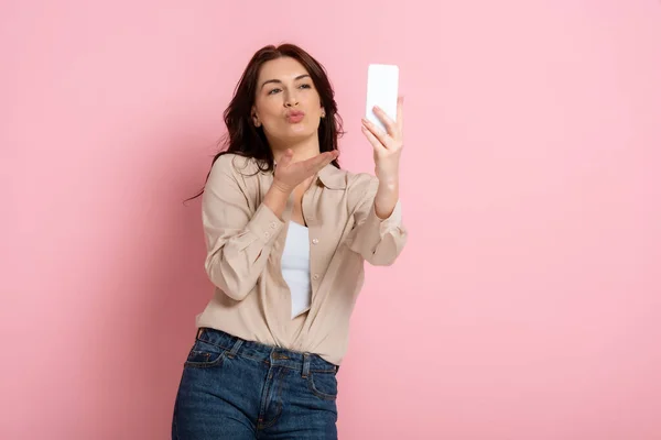 Красивая женщина дует воздушный поцелуй во время принятия селфи со смартфоном на розовом фоне, концепция тела положительный — стоковое фото