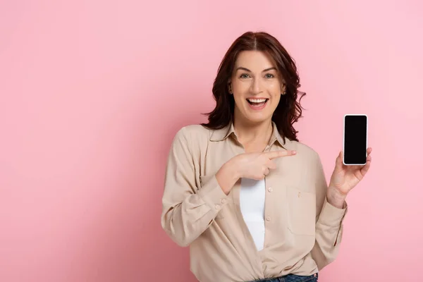 Hermosa mujer sonriendo a la cámara mientras apunta con el dedo al teléfono inteligente con pantalla en blanco sobre fondo rosa - foto de stock