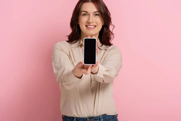 Вибірковий фокус усміхненої жінки, що показує смартфон з порожнім екраном на рожевому фоні — стокове фото