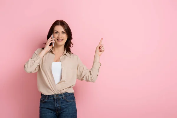 Schöne brünette Frau zeigt mit dem Finger, während sie auf dem Smartphone auf rosa Hintergrund spricht — Stockfoto