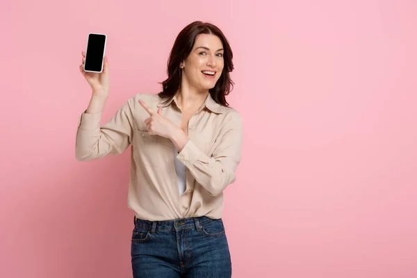 Mujer morena positiva apuntando con el dedo al smartphone con pantalla en blanco sobre fondo rosa - foto de stock