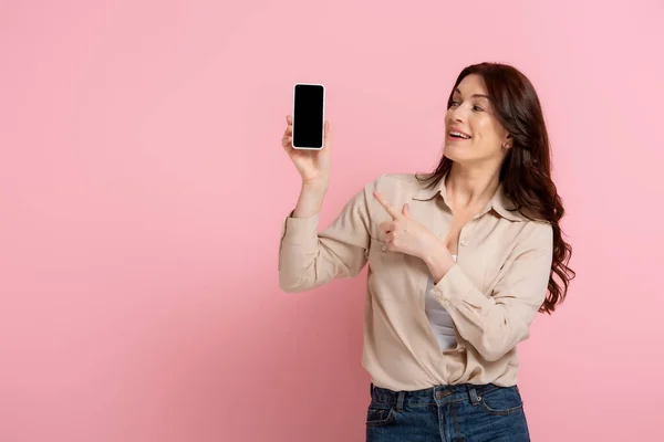 Glückliche brünette Frau zeigt mit dem Finger auf Smartphone mit leerem Bildschirm auf rosa Hintergrund — Stockfoto