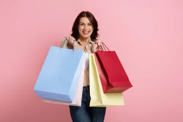 Beautiful brunette woman smiling at camera while holding colorful shopping bags on pink background — Stock Photo