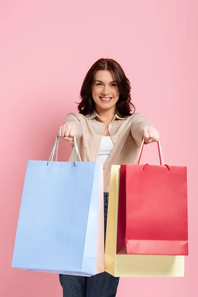Felice donna bruna sorridente alla macchina fotografica e mostrando borse della spesa su sfondo rosa — Foto stock