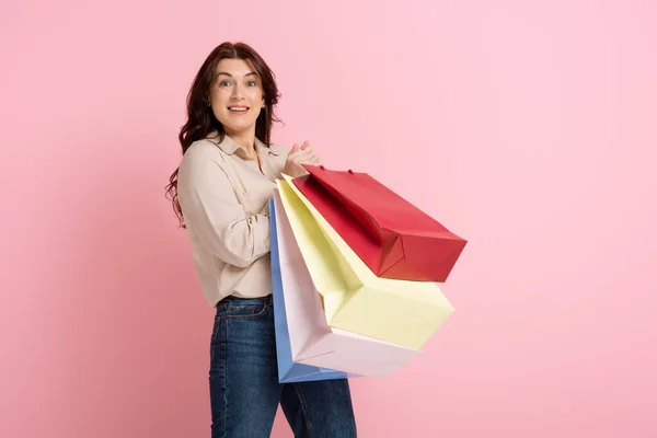 Heureuse femme brune regardant la caméra tout en tenant des sacs à provisions sur fond rose — Photo de stock