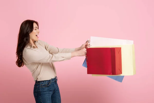 Seitenansicht einer fröhlichen Frau, die Einkaufstüten auf rosa Hintergrund hält, Konzept des positiven Körpers — Stockfoto