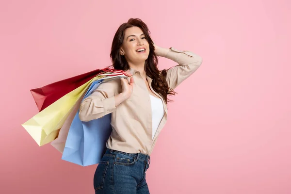 Beautiful brunette woman smiling at camera while holding colorful shopping bags on pink background — Stock Photo