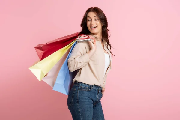 Donna positiva sorridente mentre guarda borse della spesa isolate sul rosa, concetto di corpo positivo — Foto stock