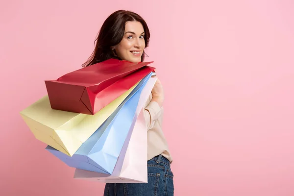 Vista posterior de la hermosa mujer sonriendo a la cámara mientras sostiene bolsas de compras aisladas en rosa - foto de stock