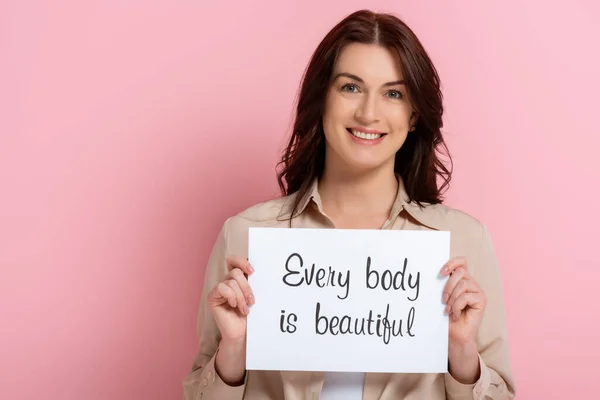 Beautiful smiling woman holding card with every body is beautiful lettering on pink background — Stock Photo