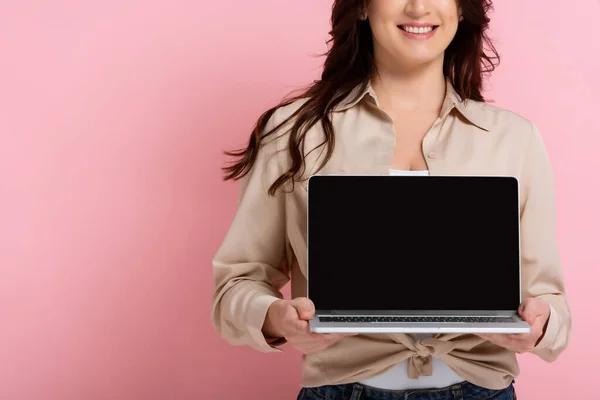 Обрезанный вид улыбающейся женщины, показывающей ноутбук с чистым экраном на розовом фоне — стоковое фото