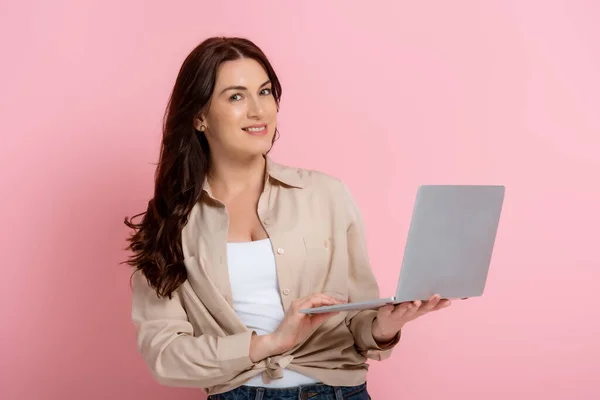 Hermosa mujer sonriente mirando a la cámara y sosteniendo el ordenador portátil sobre fondo rosa - foto de stock