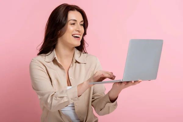 Attraktive brünette Frau lächelt, während sie Laptop auf rosa Hintergrund benutzt — Stockfoto