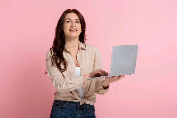 Brunette woman smiling at camera and using laptop on pink background — Stock Photo