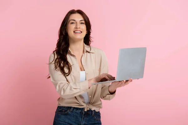 Mulher morena sorrindo para a câmera e usando laptop no fundo rosa — Fotografia de Stock