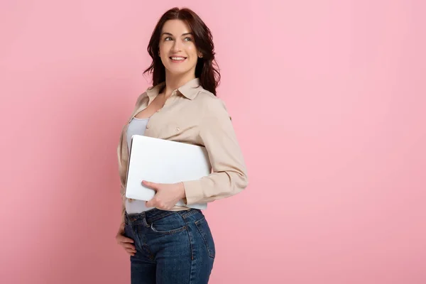 Positive Frau lächelt weg, während sie Laptop auf rosa Hintergrund hält, Konzept des positiven Körpers — Stockfoto