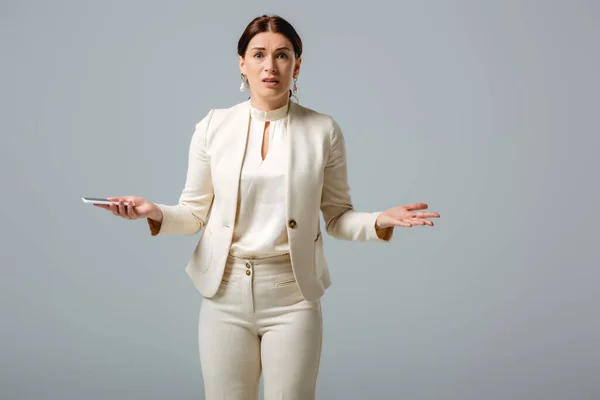 Mujer confusa en ropa formal mirando a la cámara mientras sostiene el teléfono inteligente aislado en gris, concepto de cuerpo positivo - foto de stock