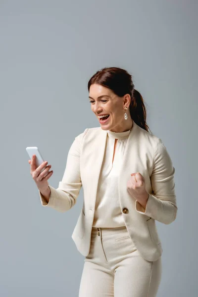 Cheerful businesswoman showing yeah gesture while holding smartphone isolated on grey — Stock Photo