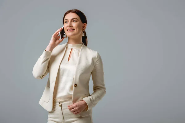 Mujer atractiva en ropa formal sonriendo mientras habla en un teléfono inteligente aislado en gris - foto de stock