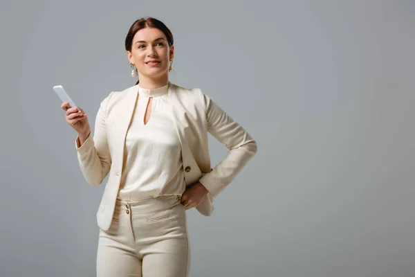 Schöne Geschäftsfrau lächelt weg, während sie ihr Smartphone isoliert auf grau hält — Stockfoto