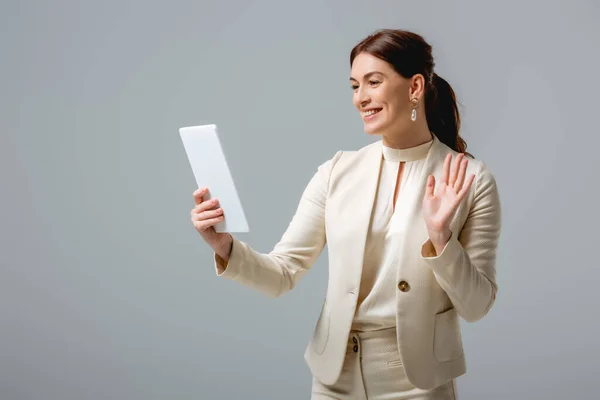 Mujer de negocios sonriente saludando de la mano mientras tiene videollamada en tableta digital aislada en gris - foto de stock