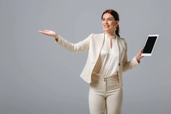 Hermosa mujer sonriente en ropa formal apuntando con la mano y sosteniendo la tableta digital con pantalla en blanco aislada en gris - foto de stock
