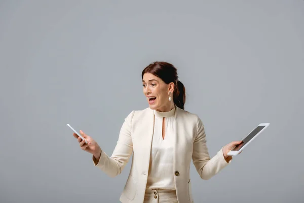 Mujer de negocios asustada sosteniendo teléfono inteligente y tableta digital aislado en gris - foto de stock