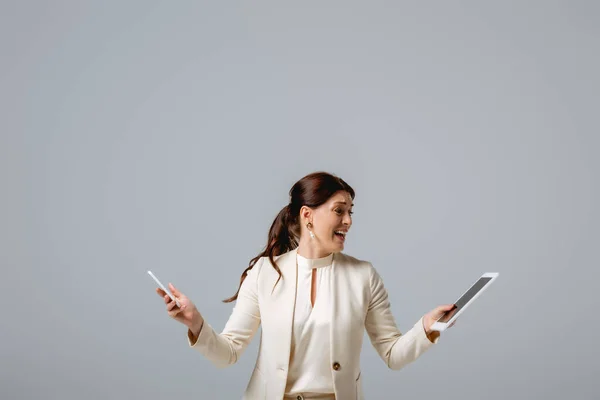 Налякана жінка в офіційному одязі тримає цифровий планшет і мобільний телефон ізольовані на сірому — стокове фото