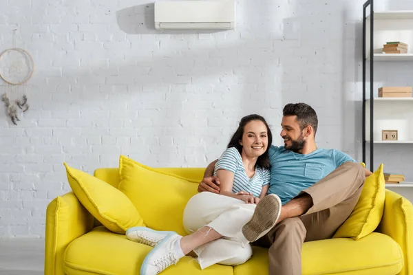 Guapo barbudo hombre abrazando sonriente novia en sofá en sala de estar - foto de stock