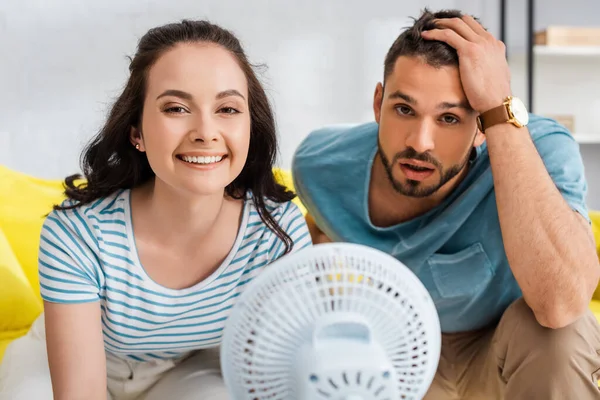 Foco seletivo de menina sorridente olhando para a câmera perto de namorado cansado e ventilador elétrico — Fotografia de Stock