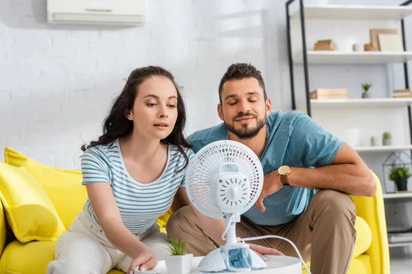 Pareja joven sentada cerca de ventilador eléctrico en la mesa de café en la sala de estar - foto de stock