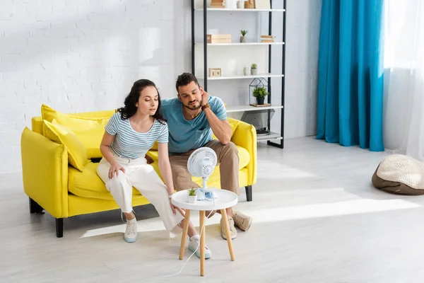 Junges Paar blickt auf elektrischen Ventilator auf Couchtisch, während es auf Sofa im Wohnzimmer sitzt — Stockfoto