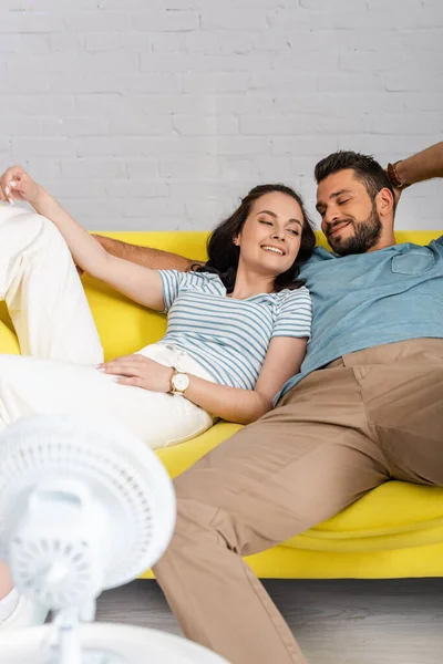 Selektiver Fokus einer jungen Frau, die neben ihrem lächelnden Freund auf der Couch neben einem Elektroventilator liegt — Stockfoto