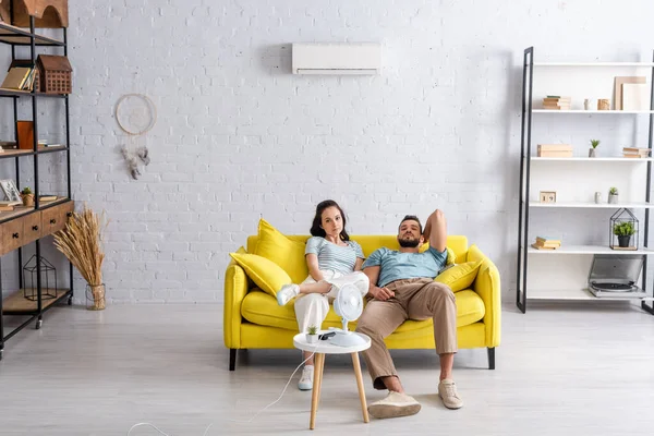 Молодая пара смотрит в камеру, сидя на диване рядом с электрическим вентилятором и кондиционером на стене в гостиной — стоковое фото