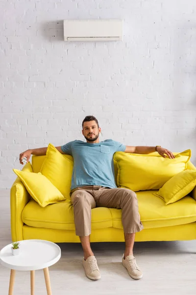 Selektiver Fokus eines gutaussehenden Mannes mit Fernbedienung der Klimaanlage, während er zu Hause auf dem Sofa in die Kamera schaut — Stockfoto